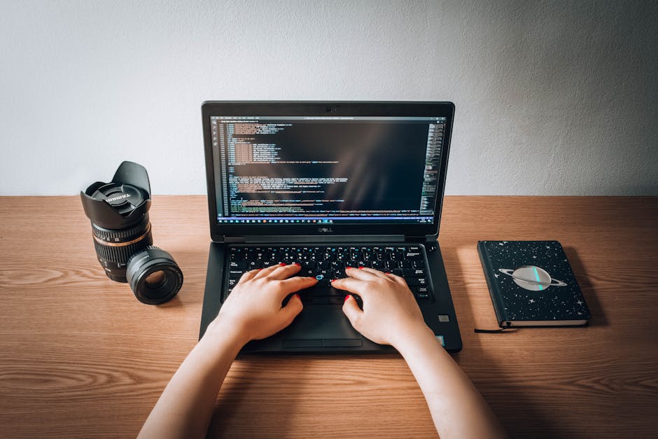 A top-down view of a wooden desk with a person's hands typing on a black Dell laptop, which displays lines of code on its screen. To the left of the laptop, there is a black DSLR camera with a large l