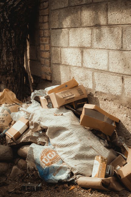 A person's hand wearing a pink rubber glove is holding a clear plastic bag filled with crumpled and flat plastic bottles, representing collected recyclable waste. The background is a plain yellow curt