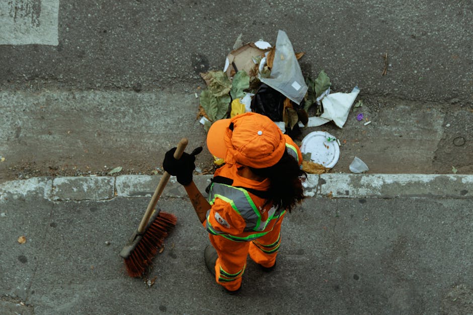 A sanitation worker in a high-visibility orange vest is standing at the rear of a large red rubbish collection truck on a street. The worker is engaged in emptying or maintaining the truck, which has